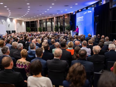 Eine Frau steht auf einer Bühne und hält ein Vortrag, davor sitzt ein Publikum