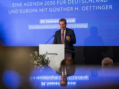 älterer Mann steht auf einer Bühne an einem Mikrofon, davor sitzt ein Publikum 