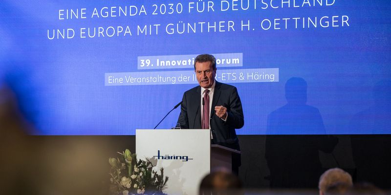 älterer Mann steht auf einer Bühne an einem Mikrofon, davor sitzt ein Publikum 